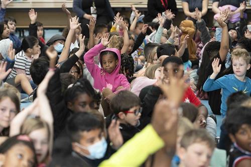 A crowded classroom with about 250 students sitting together. One student has her hand raised.